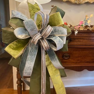 May include: A large decorative bow with multiple layers of green, olive, and silver-blue velvet ribbons. The bow is accented with a central section of shimmering, light brown ribbon with silver trim. The bow is attached to a lamp.