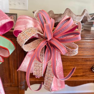 May include: A large decorative bow with multiple layers of pink velvet and woven ribbon. The bow features a central knot with long, flowing tails. The color palette includes shades of pink, coral, and a touch of woven beige.