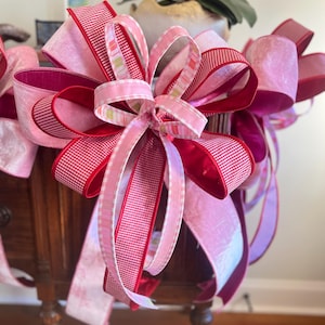 May include: A decorative bow featuring layers of pink and red ribbons. The bow has a multi-layered design with a mix of solid, striped, and patterned ribbons. The ribbons are arranged to create a full, voluminous shape, suitable for gift wrapping or holiday decor.