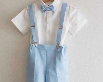 May include: A white linen shirt with short sleeves and a light blue bow tie. The shirt has a button-up front and is paired with light blue linen shorts and suspenders.