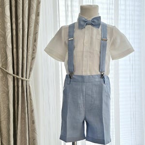 May include: A white linen shirt with short sleeves and a light blue bow tie. The shirt has suspenders attached and is worn over a pair of light blue linen shorts.