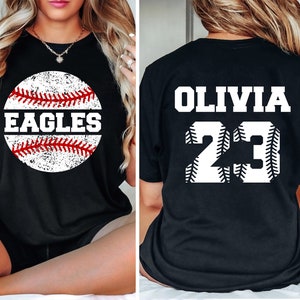 May include: Black t-shirt with a white baseball graphic. The baseball has red stitching and the word "EAGLES" written on it. The back of the shirt has the name "OLIVIA" and the number "23" in white with red stitching.