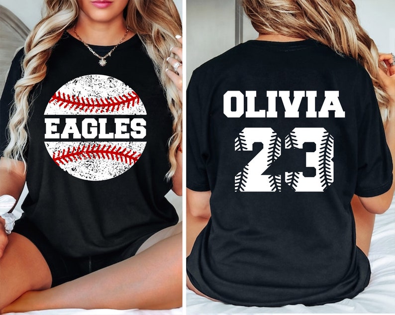 May include: Black t-shirt with a white baseball graphic. The baseball has red stitching and the word "EAGLES" written on it. The back of the shirt has the name "OLIVIA" and the number "23" in white with red stitching.