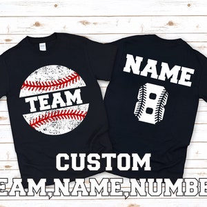 May include: Two black t-shirts with a baseball theme. The front of one shirt has a white baseball with red stitching and the word "TEAM" in red. The back of the other shirt has a white baseball-shaped number "8" and the word "NAME" in white.