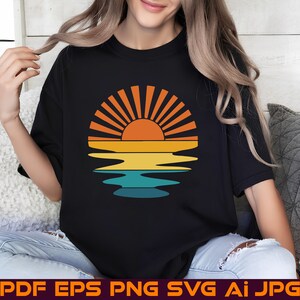 以下が含まれることがあります： レトロなスタイルの夕焼けのグラフィックがプリントされた黒いTシャツ。太陽はオレンジとイエローで、そこから光線が伸びています。太陽は下にある水に反射しており、水は青と緑です。