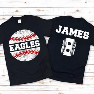 May include: Two black t-shirts with a baseball theme. One shirt has a white baseball with red stitching and the word "EAGLES" on it. The other shirt has the name "JAMES" and the number "8" in white with baseball stitching.
