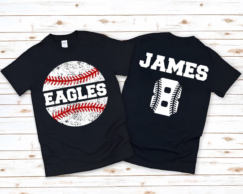 May include: Two black t-shirts with a baseball theme. One shirt has a white baseball with red stitching and the word "EAGLES" on it. The other shirt has the name "JAMES" and the number "8" in white with baseball stitching.