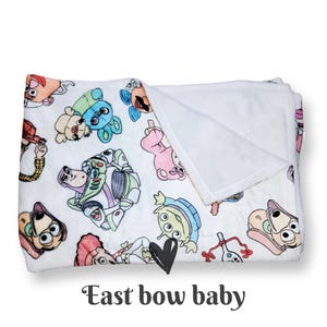 Może przedstawiać: Biały pluszowy koc z kolorowym nadrukiem postaci z kresk&oacute;wek z serii Toy Story. Koc ma białe polarowe podszewka.