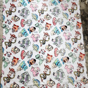 Może przedstawiać: Biały koc z polaru z powtarzającym się wzorem postaci z Toy Story, w tym Woody, Buzz Astral, Pan Potato Head i Rex. Postacie są przedstawione w kresk&oacute;wkowym stylu i otoczone białym tłem.