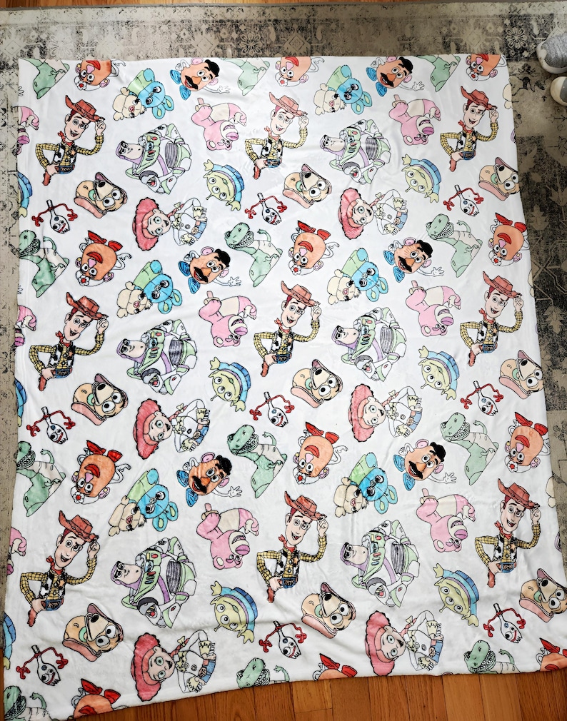 Może przedstawiać: Biały koc z polaru z powtarzającym się wzorem postaci z Toy Story, w tym Woody, Buzz Astral, Pan Potato Head i Rex. Postacie są przedstawione w kresk&oacute;wkowym stylu i otoczone białym tłem.