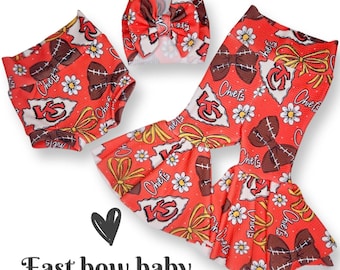 May include: A red, white, and brown baby outfit with a football and bow pattern. The outfit includes a headband, a pair of bloomers, and a pair of bell bottoms. The outfit has the text "Chiefs" on it.