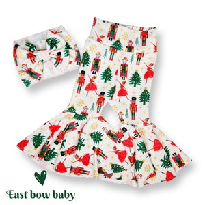 May include: White bell bottom pants with a Nutcracker and Christmas tree print. The pants have a wide leg opening and a small slit in the front. A white headband with a red bow is included.