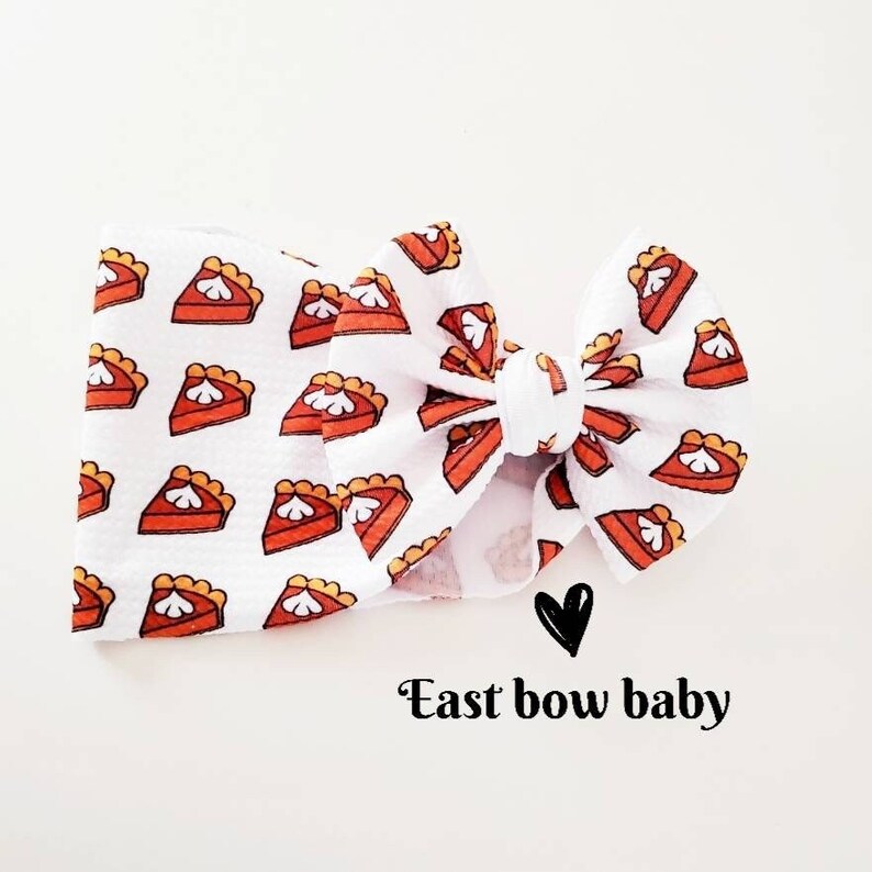 May include: White fabric headband with a repeating pattern of cartoon pie slices in orange, brown, and cream. The headband is tied in a bow. The text "East bow baby" is below the bow.