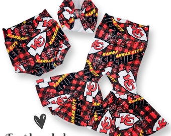 May include: A baby clothing set featuring a red, black, and white Kansas City Chiefs design. The set includes flared pants, a headband, and a diaper cover. The words "Kansas City Chiefs" are visible. The text "East bow baby" is also visible.