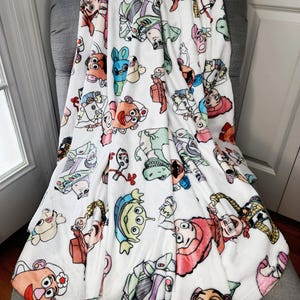 Może przedstawiać: Biały pluszowy koc z kolorowym wzorem postaci z Toy Story, w tym Woody, Buzz Astral i Pan Potato.