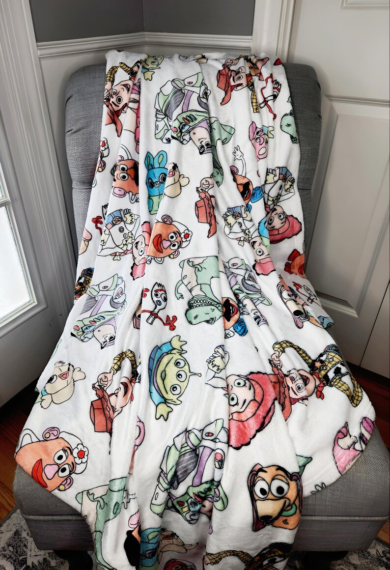 Może przedstawiać: Biały pluszowy koc z kolorowym wzorem postaci z Toy Story, w tym Woody, Buzz Astral i Pan Potato.