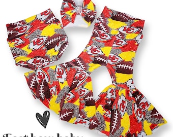 May include: A baby outfit set featuring a red, yellow and brown patterned skirt with a matching headband. The skirt has a ruffled hem and the pattern includes footballs, the letter "K" and the number "15". The headband has a white bow with a rhinestone in the centre. The text "East bow baby" is visible below the outfit.