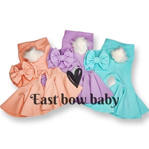 May include: Three pairs of baby leggings with matching bows in peach, lavender, and turquoise. Each pair has a fluffy white pom-pom on the back. The text "East bow baby" is printed on the image.