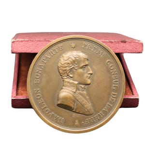 Op de afbeelding: Een bronzen medaille met een profiel van Napoleon Bonaparte, met Franse tekst. De medaille wordt gepresenteerd in een rode presentatiedoos met een fluwelen binnenkant. De ronde medaille heeft een gedetailleerd reliëf.