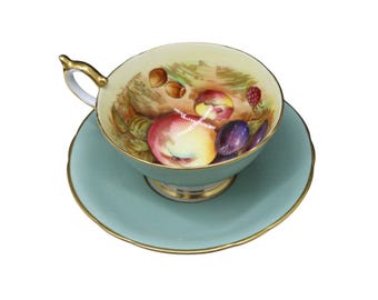 May include: A vintage teacup and saucer set. The cup and saucer are a light teal color with gold trim. The inside of the cup features a hand-painted design of peaches, plums, and hazelnuts. The handle of the cup is gold.