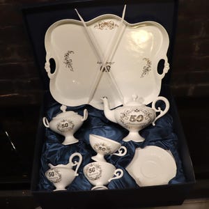 May include: A white porcelain tea set, including a tray, teapot, sugar bowl, creamer, cup, and saucer. Each piece features a black floral design and the number "50" in a wreath, possibly for an anniversary. The set is presented in a blue box.