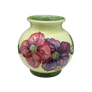 May include: A ceramic vase with a rounded body, green rim and base. The vase features two large, stylised flowers in red and purple, with green centres, on a pale yellow background. A decorative item.