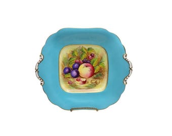 May include: A square, turquoise-colored serving dish with a scalloped edge and gold trim. The center features a painted still life of fruit, including plums, peaches, and berries. The dish has decorative handles on either side.