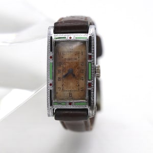 May include: A rectangular Bulova wristwatch with a brown leather strap. The silver-tone metal case features black and green enamel accents with small red diamond details. The watch face is aged tan with black Arabic numerals and two hands.