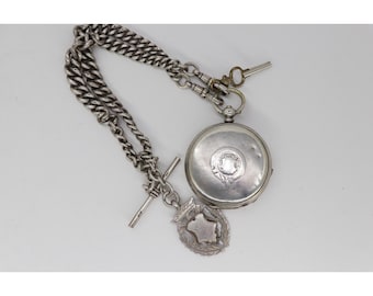 Könnte beinhalten: Eine silberne Taschenuhr mit Kette und Schlüssel. Die Uhr hat ein rundes Zifferblatt mit einem dekorativen Design. An der Uhr ist eine silberne Kette befestigt, zusammen mit einem dekorativen Anhänger. Die Uhr und die Kette befinden sich auf weißem Hintergrund.