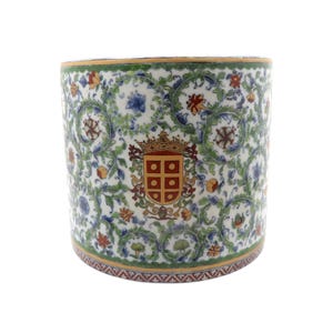 Puede incluir: Un recipiente cilíndrico de porcelana con fondo blanco y un intrincado patrón de enredaderas y flores verdes. Un escudo heráldico central en rojo y oro, rematado con una corona, se muestra de forma prominente en la parte frontal del objeto.