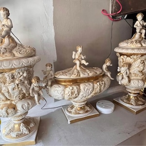 Könnte beinhalten: Verzierte cremefarbene Keramikurnen mit Goldakzenten und Cherubfiguren. Die Urnen weisen detaillierte Reliefschnitzereien und Deckel mit Cherubskulpturen auf. Die Sammlung umfasst zwei große Urnen und ein kleineres, verziertes Mittelstück.