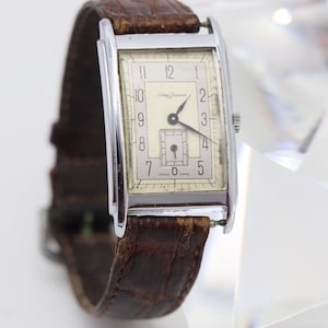 May include: A rectangular wristwatch with a silver-tone metal case and a brown textured leather strap. The off-white dial features black Arabic numerals, a small sub-dial at the six o'clock position, black hands, and the text Swiss Made.