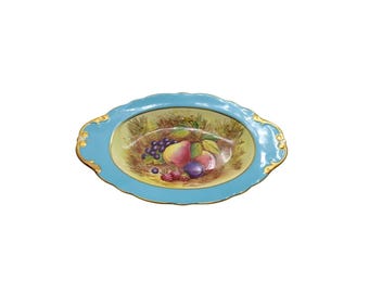 May include: Oval-shaped ceramic serving dish with a light blue border and gold trim. The center features a painted still life of fruit, including grapes, pears, and plums. A decorative piece for serving or display.