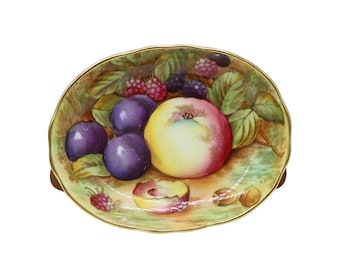May include: A decorative, oval-shaped plate with a painted still life of fruit. The plate features an apple, plums, raspberries, and blackberries. The plate has a gold rim and a green and yellow background.