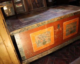 May include: Antique wooden chest with a red painted exterior and decorative floral panels. The chest has a rectangular shape with a flat top and is likely used for storage. The chest has a distressed appearance, suggesting age and use.