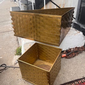 May include: Two gold-colored metal planters with a woven basket design. One is rectangular, the other is square. The planters are sitting on a concrete surface, suggesting outdoor use. The design gives a vintage or antique feel.