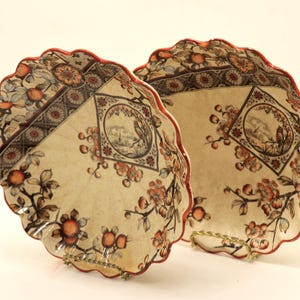 Peut inclure: Deux assiettes décoratives avec un motif floral et des bords festonnés. Les assiettes ont un fond crème avec des motifs floraux orange et marron et une bordure rouge. Chaque assiette présente un paysage unique.