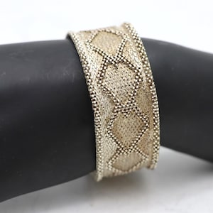 May include: A wide, silver-toned bracelet with a woven design featuring diamond-shaped patterns. The bracelet is displayed on a black surface, showcasing its intricate texture and metallic sheen. A piece of jewelry.