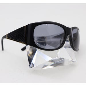 Puede incluir: Gafas de sol negras con lentes gris oscuro y una montura gruesa de color negro brillante. Las patillas muestran un patrón texturizado similar a la piel de serpiente en negro con finos detalles dorados. El nombre Jimmy Choo aparece impreso en oro.