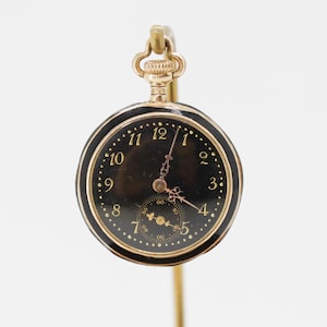 Peut inclure: Une montre de poche dorée avec un cadran noir et des chiffres dorés. La montre a un petit cadran de secondes et est suspendue à un crochet doré. Les aiguilles de la montre sont dorées.
