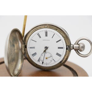 May include: A silver pocket watch with an open face, revealing a white dial with black Roman numerals and the brand name "LONGINES". The watch has a small seconds dial and a winding stem. The watch is displayed on a wooden surface.