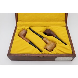 May include: A wooden presentation box with a yellow velvet interior containing three tobacco pipes. The pipes feature smooth and textured brown wooden bowls with black stems. A metal plaque on the front of the box displays engraved text.