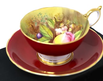 Può includere: Un set da tè in rosso e oro con un design floreale che presenta frutta e foglie. La tazza e il piattino sono decorati con un bordo dorato.