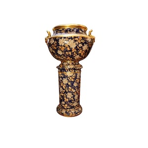 Pode incluir: Um vaso decorativo e ornamentado com fundo azul escuro e padrão floral dourado. O vaso tem um topo largo e arredondado com alças e acabamentos dourados, e assenta sobre uma coluna com um design correspondente.