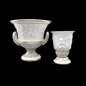 May include: Two white ceramic vases with gold detailing. The larger vase has handles and a detailed relief of figures. The smaller vase features a relief of cherubs. Both vases have a pedestal base and are set against a black background.