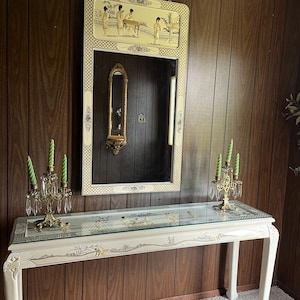 May include: A white console table with a glass top and ornate carvings. The table has a painted design of flowers and birds. The table is in front of a brown wood wall with a large mirror above it. The mirror has a gold frame and a painted scene of people in a garden.