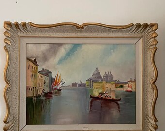 Puede incluir: Pintura al óleo de una escena de canal veneciano en un marco decorativo. La obra representa edificios, barcos y una iglesia con cúpula bajo un cielo nublado. El marco es blanco con detalles dorados.