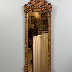 May include: Ornate, antique, rectangular mirror with a carved wooden frame. The frame is a warm, golden brown color and features intricate floral and scrollwork designs. The top of the mirror has a decorative oval element with a bow. The mirror reflects the room.