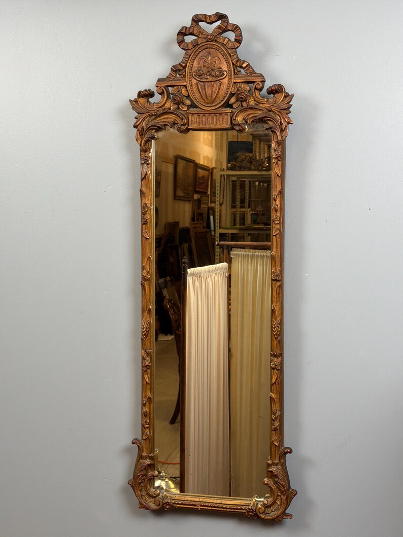 May include: Ornate, antique, rectangular mirror with a carved wooden frame. The frame is a warm, golden brown color and features intricate floral and scrollwork designs. The top of the mirror has a decorative oval element with a bow. The mirror reflects the room.