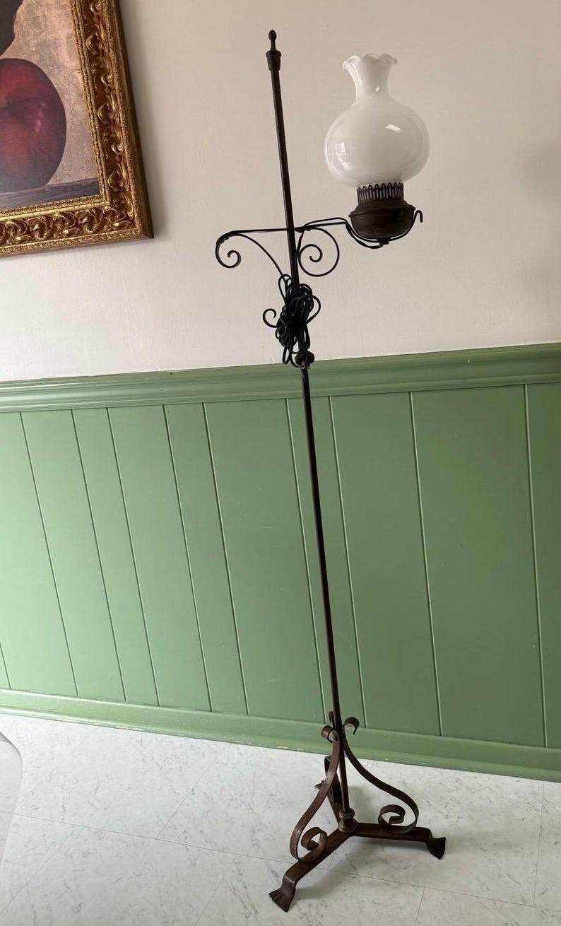 May include: An antique floor lamp with a dark metal frame and a white glass globe shade. The lamp features decorative scrollwork and a slender, vertical design. The base has a three-footed design. The lamp is a vintage lighting fixture.